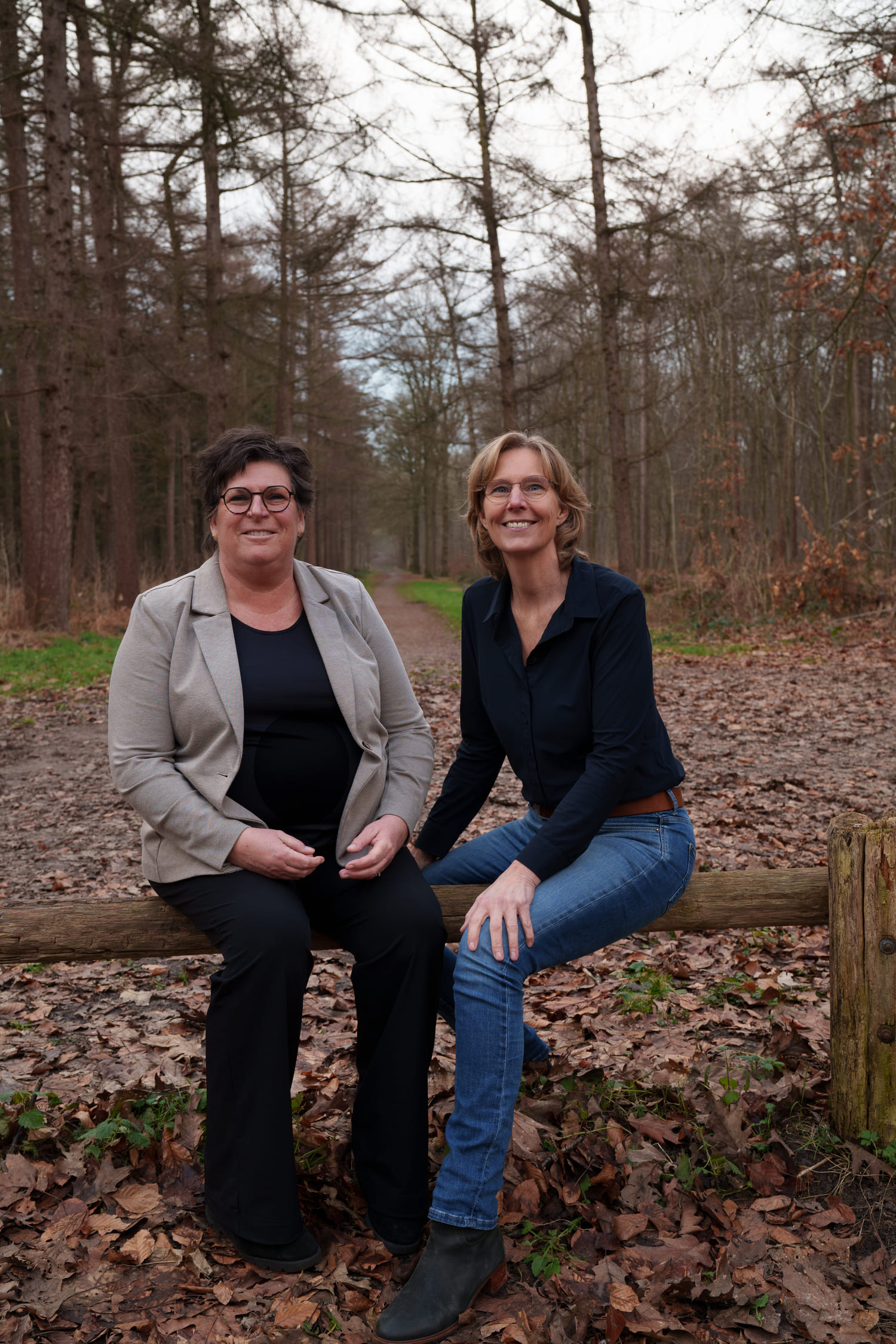 Twee vrouwen zittend op een houten hek in een bos, genietend van een ontspannen en vriendelijke sfeer – Manuele Therapie Prinsenbeek.